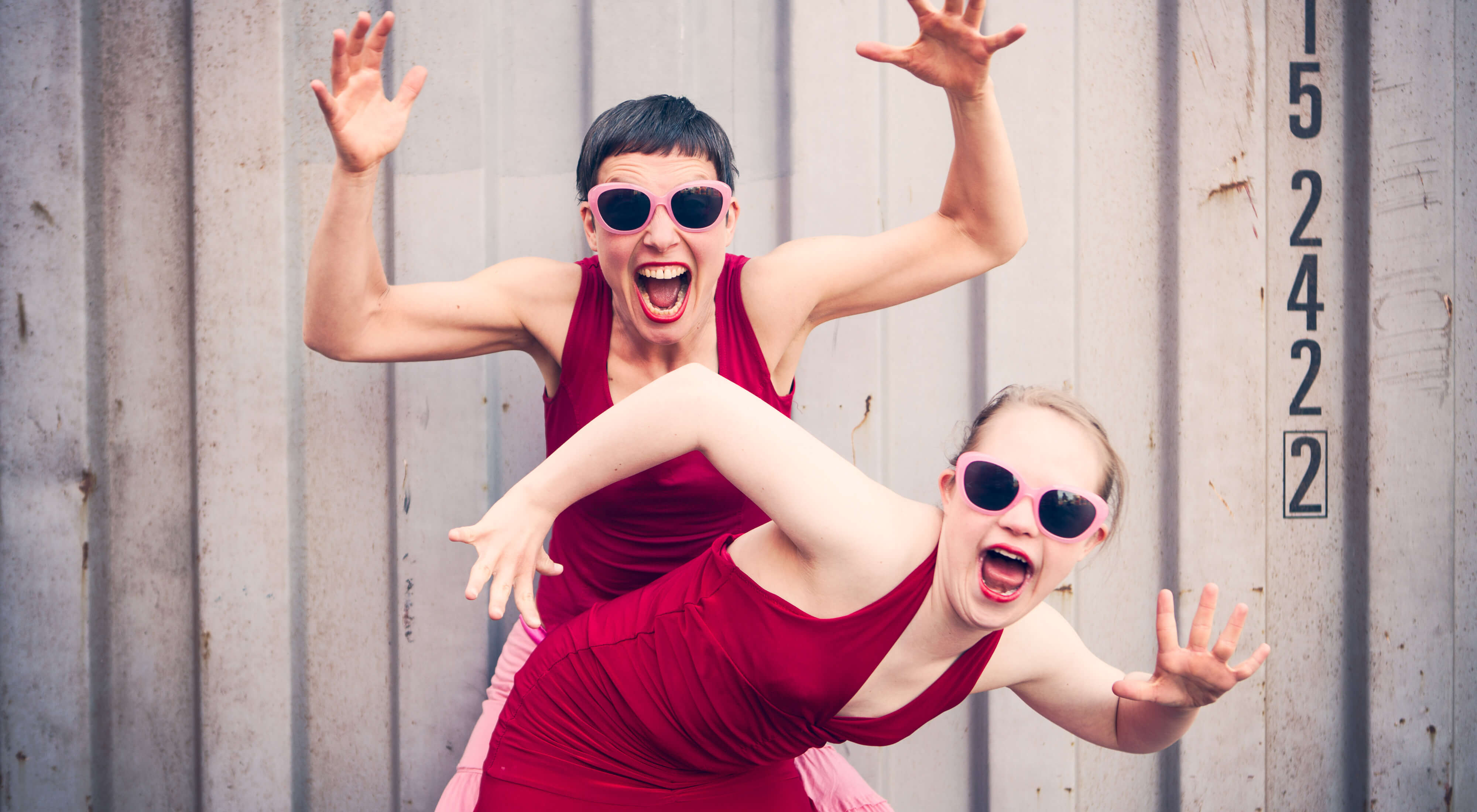 Theaterbild: Zwei junge Frauen in dunkelroten Sommerkleidern halten die Arme hoch und schreien, eine davon mit Downsyndrom