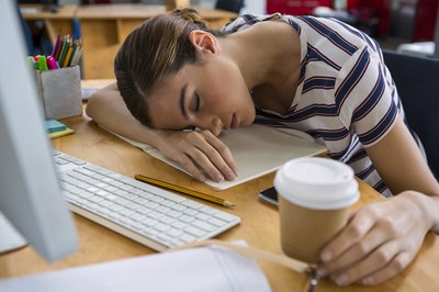 Eine junge Frau schläft mit dem Kopf auf dem Schreibtisch, die linke Hand noch am Kaffeebecher.  