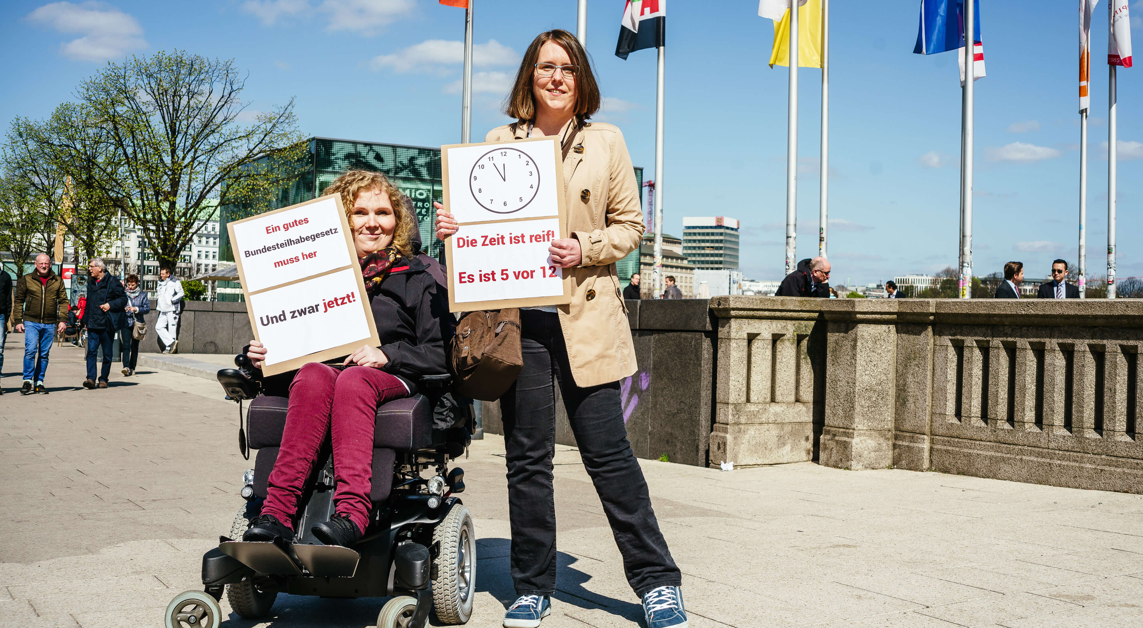 Zwei Frauen halten Protestschilder hoch für "Teilhabe Jetzt". Eine Frau sitzt im Rollstuhl. Sie sind auf einer Brücke in Hamburg, Fahnen im Hintergrund.